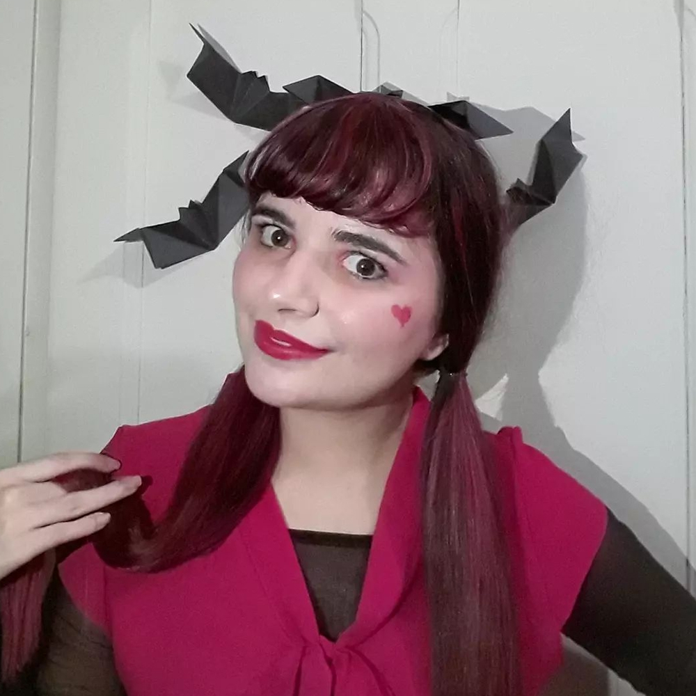 Foto de Luly Lage com o cabelo preso em marias chiquinhas vestindo uma blusa de frio fina preta por baico de uma rosa com gola que termina em um laço, maquiagem completamente rosa com um coração desenhado na bochecha esquerda. Ao fundo, alguns morcegos de papel foram fixados à porta branca e há um preso em seus cabelos.