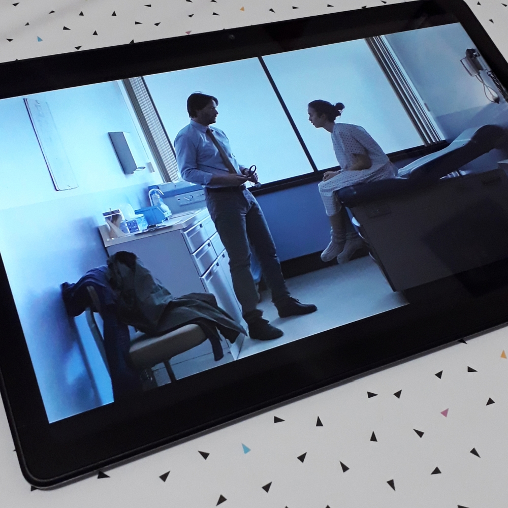 Foto de um tablet onde se vê uma cena do filme To The Bone: O Minimo Para Viver, representando uma consulta do médico interpretado por Keanu Reeves conversando com sua paciente, interpretada por Lily Collins, sentada em uma maca de frente para ele.