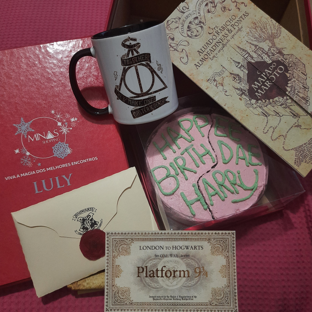 Itens inspirados na série Harry Potter sobre um fundo cor de rosa: carta de Hogwarts em envelope bege com brasão da escola e lacre em cera vermelho, caneca com o símbolo das Relíquias da Morte, bolo cor de rosa onde se lê 'Hapee birthday Harry' em verde, Mapa do Maroto e uma passagem para o embarque na plataforma rumo à escola, todos decorativos à exceção do bolo e da caneca.