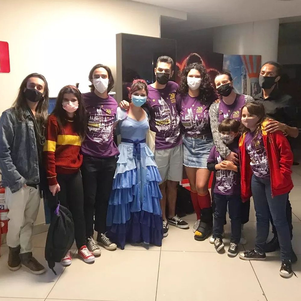 Grupo de 10 pessoas, lado a lado posando para a foto, todas elas usando máscaras de proteção. São, ao todo, 4 homens, 4 mulheres, um menino e uma menina. Eles estão localizados na saída do cinema de um shopping.