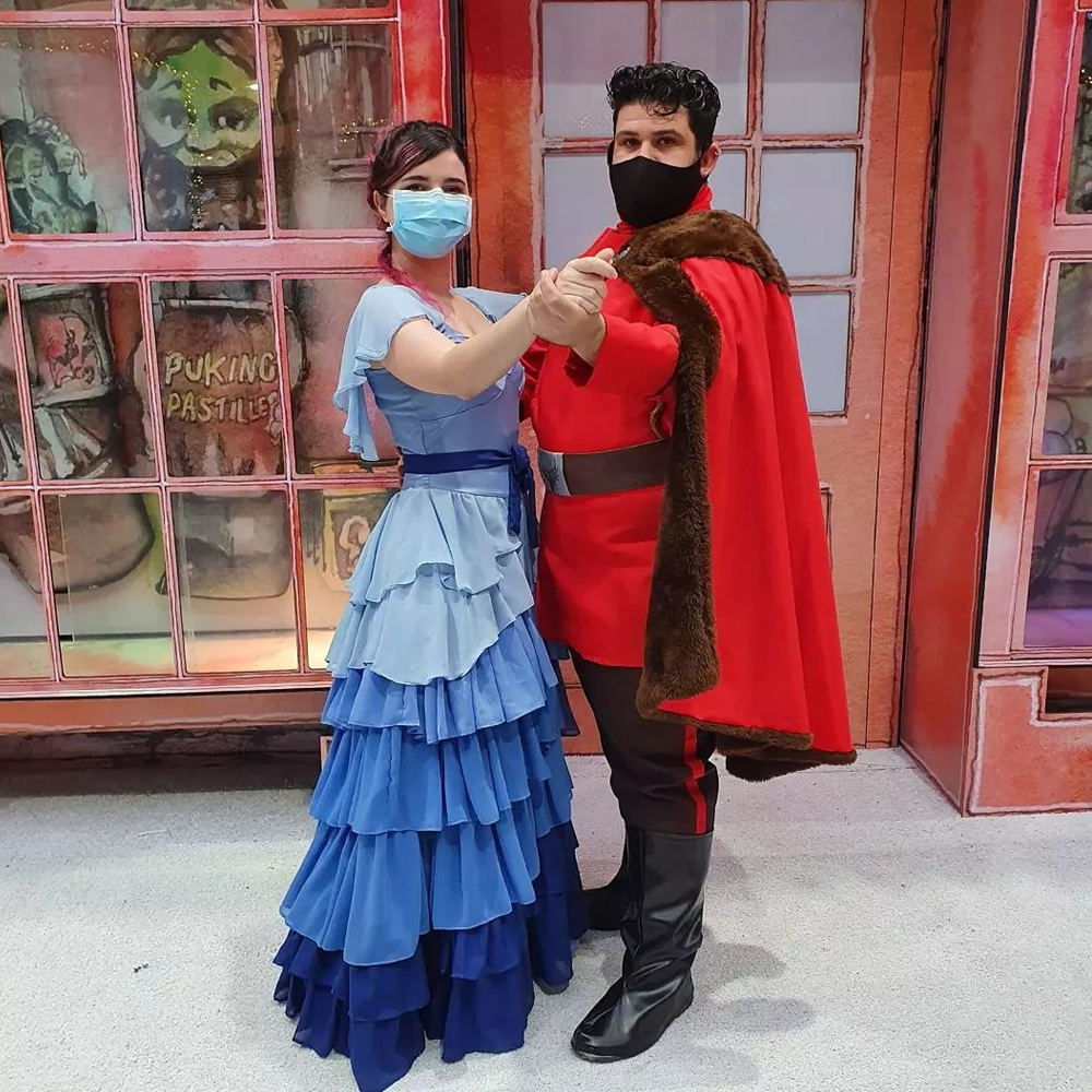 Foto de uma mulher branca usando um vestido de baile azul, com a saia em degradê indo do claro para o escuro, inspirado no vestido da personagem Hermione, com cabelos cor de rosa e pretos preso e em coque usando máscara azul sobre nariz e boca, que parece valsar com um homem branco de cabelos escuros usando capa vermelha inspirada na do personagem Victor Krum. Os dois estão em um cenário de natal baseado na série Harry Potter, de onde seus trajes foram tirados.