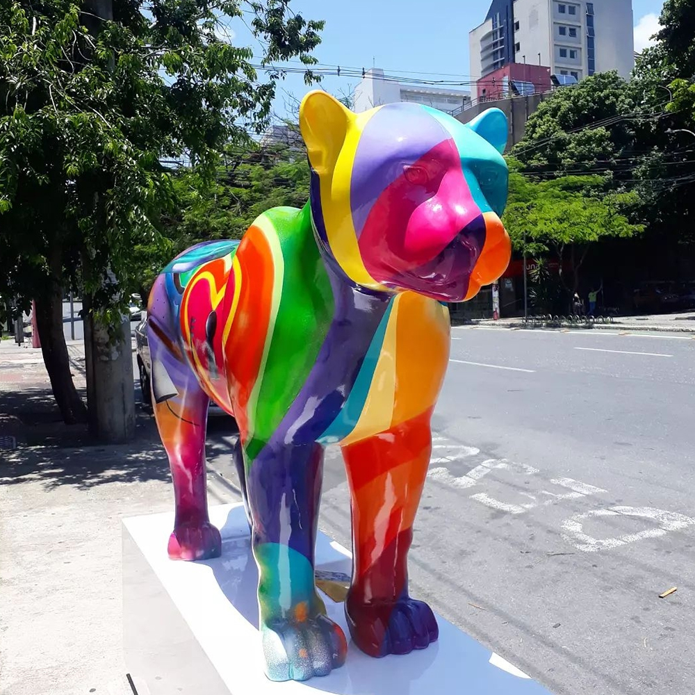 Escultura de jaguar pintada de várias cores om forma geométricas variadas, sobre um pedestal na calçada de uma avenida de Belo Horizonte.