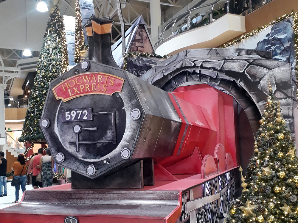 Vista da vila decorativa em close na grande locomotiva vermelha representando o Expresso de Hogwarts, cercada de árvores de Natal.