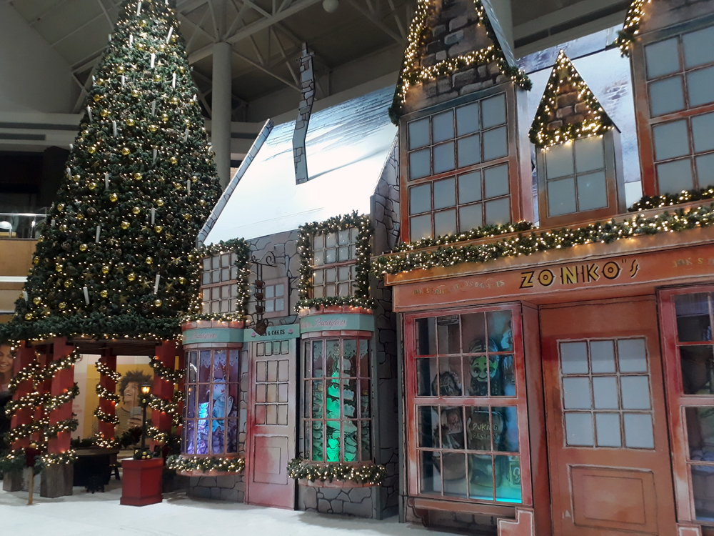 Vista da vila decorativa onde estão visíveis uma grande árvore de Natal que há sobre uma área de interação para crianças e as réplicas das lojas Casa de Chá e Zonko's