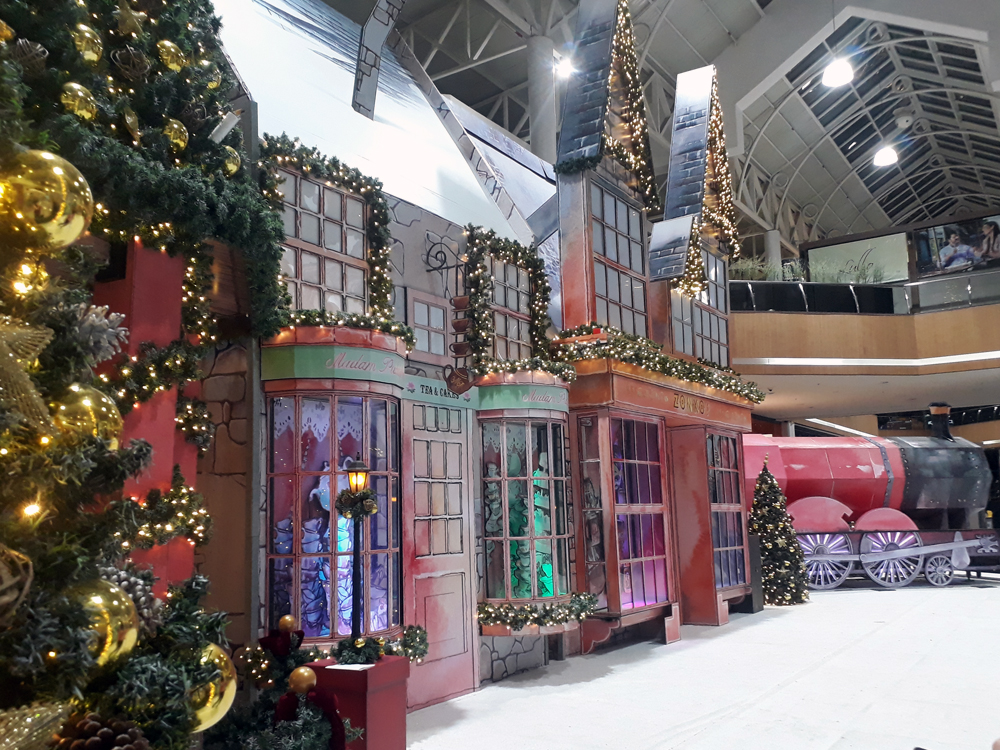 Vista da vila decorativa onde estão visíveis enfeites de Natal, as réplicas das lojas Casa de Chá e Zonko's, além do Expresso de Hogwarts ao fundo