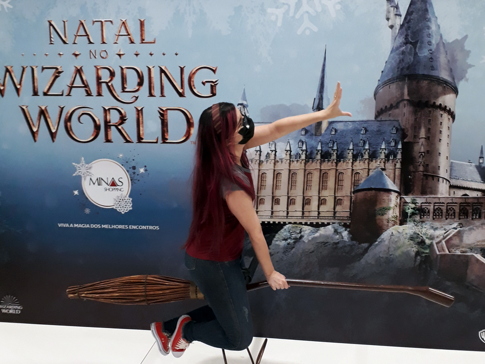Foto onde estou em um cenário onde há o castelo de Hogwarts ao fundo, onde há uma vassoura de corrida em tamanho real, na qual me encontro montada simulando pegar um pomo de ouro no alto.