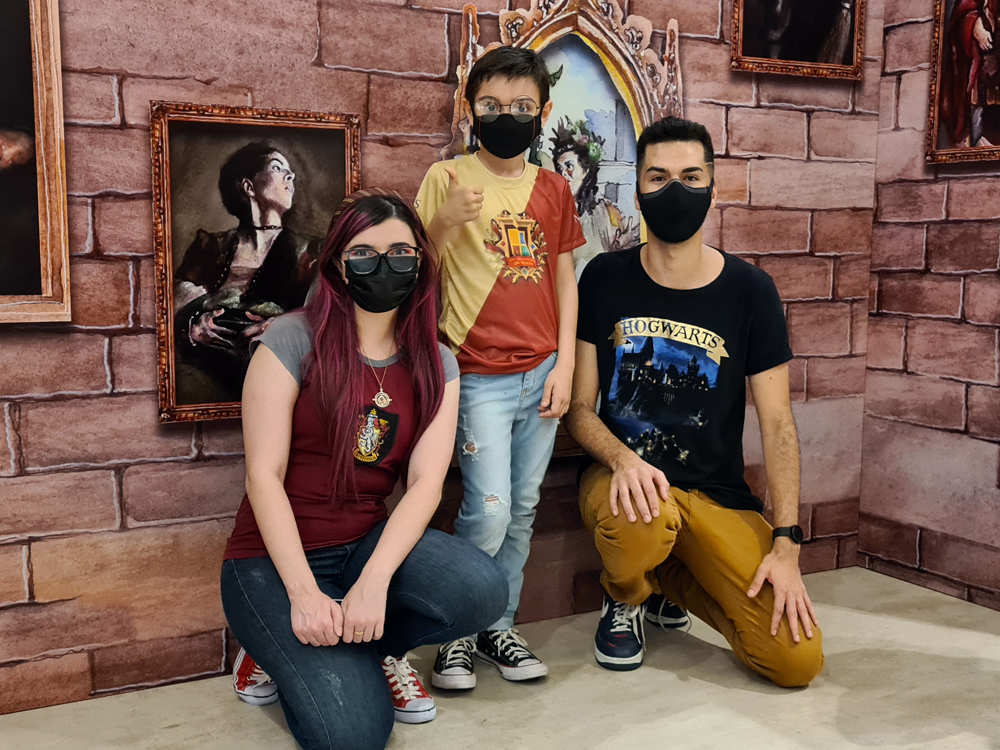 Foto onde estou ajoelhada ao lado de Gabriel Rocha, um garoto de 10 anos vestindo blusa bicolor do evento realizado no Minas Shopping e calça jeans, e Gil, com blusa escura do castelo de Hogwarts e calça mostarda, também ajoelhado. Os três estão usando máscaras de proteção pretas.