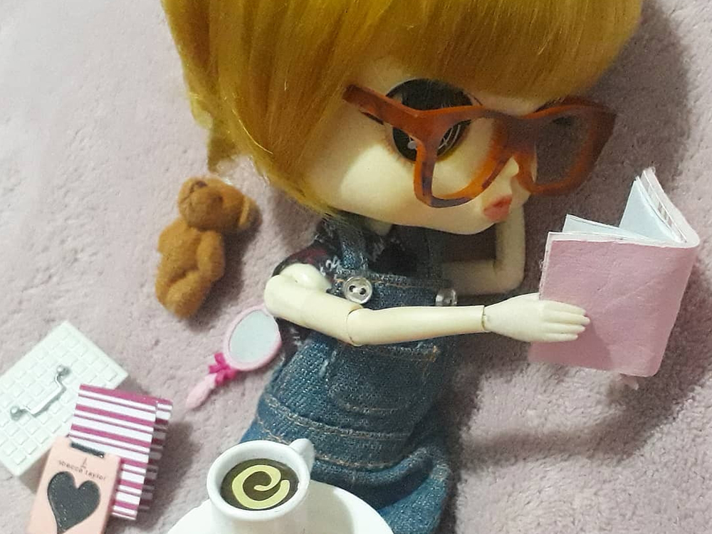 Foto da boneca Joanne referente ao trecho Mas ainda posso chegar lá do texto, agora vista na posição normal, recostada no fundo rosa lendo um caderno d ecapa da mesma cor, com o chocolate quente no colo. Ao seu lado há outros cadernos, um urso de pelúcia e um espelho de mão rosa.