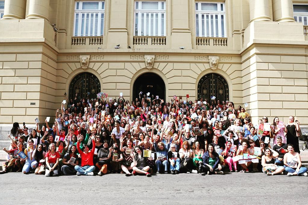 Foto da fachada do Centro Cultural Banco do Brasil Belo Horizonte, um prédio de cor clara com imponentes portas altas de topo arredondado, onde mais de 200 mulheres celebrando Um Grande Dia Para Escritoras estão posicionadas nas escadarias, muitas delas segurando seus livros e/ou com mãos levantadas em comemoração..