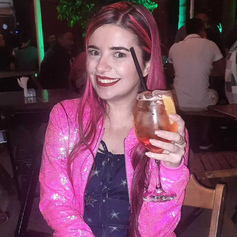 Foto minha sorridente, com a mesma roupa da foto anterior mas um pouco mais longe, segurando um drink colorido em direção à câmera.