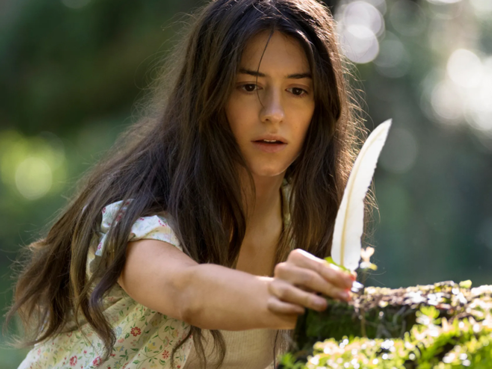 Imagem onde a protagonista do filme, uma jovem branca de longos cabelos escuros, aparece em destaque coletando uma pena de ave branca.