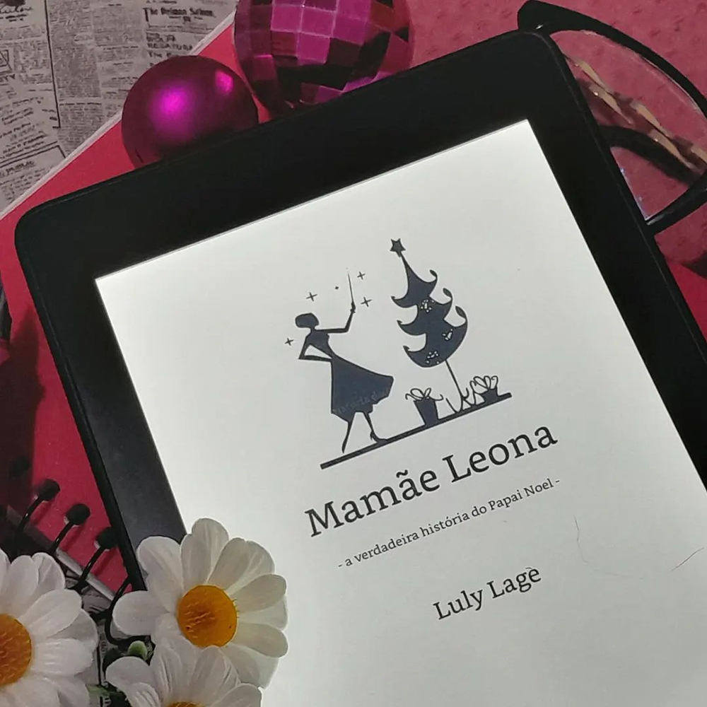 Aparelho Kindle aberto na folha de rosto do livro Mamãe Leona, uma versão simplificada da capa, sobre fundo rosa, preto e branco. Ao seu lado, nas margens da foto, margaridas falsas, bolas de natal cor de rosa, uma caneta tinteiro da memsa cor e uma armação de óculos preta.