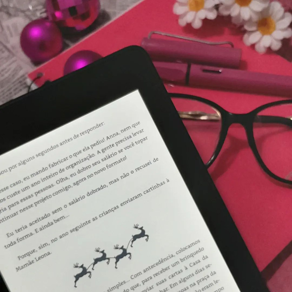 Aparelho Kindle aberto em uma das páginas do livro sobre fundo rosa, preto e branco. Ao seu lado, nas margens da foto, margaridas falsas, bolas de natal cor de rosa, uma caneta tinteiro da memsa cor e uma armação de óculos preta.