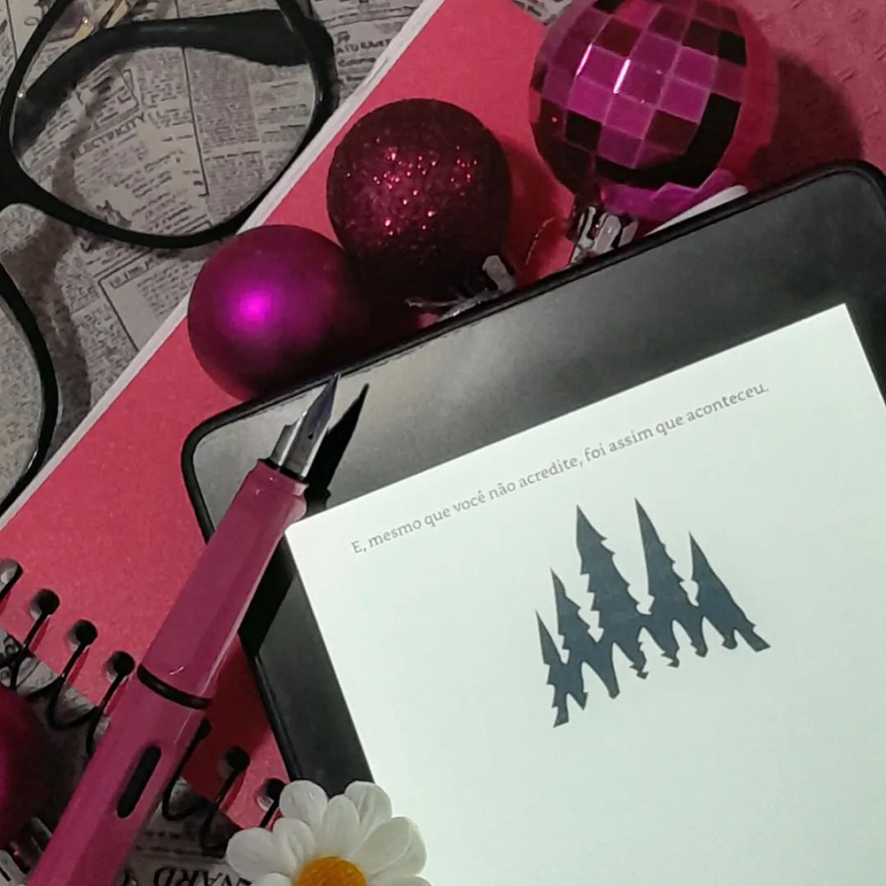 Aparelho Kindle aberto na última página do livro sobre fundo rosa, preto e branco. Ao seu lado, nas margens da foto, margaridas falsas, bolas de natal cor de rosa, uma caneta tinteiro da memsa cor e uma armação de óculos preta.