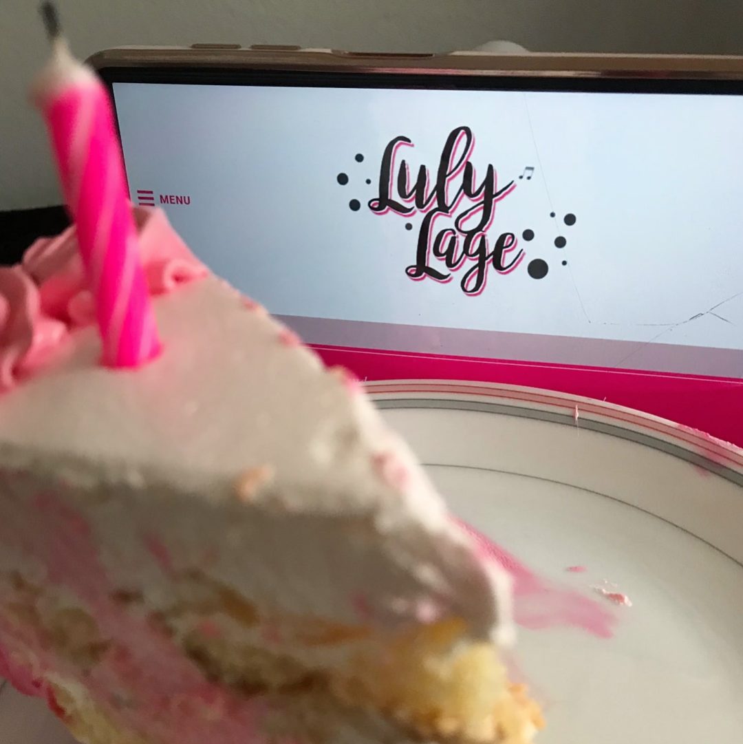 Foto de um pedaço de bolo de cobertura branca e rosa em primeiro plano, desfocado, com uma vela rosa em cima. Ao fundo, em foco, um celular deitado com a página inicial do blog aberta.