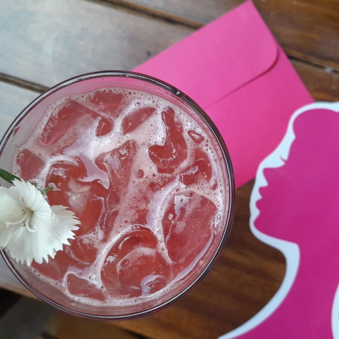 Foto de um drink rosa, visto de cima, ao lado de um envelope rosa e um display da logo da boneca Barbie, seu rosto em perfil, da mesma cor.