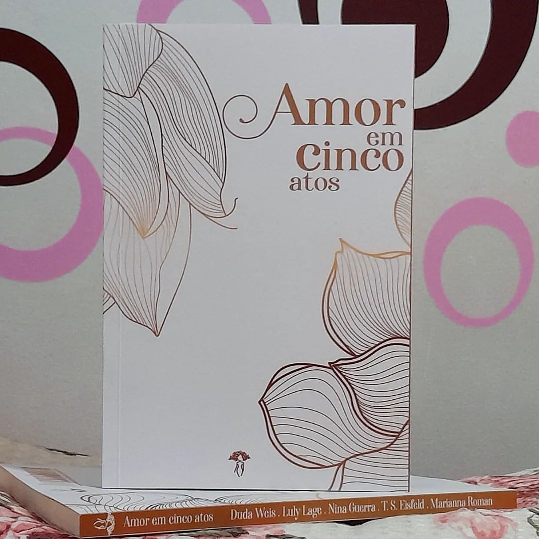 Foto do livro AMOR EM CINCO ATOS, com fundo claro e detalhes em dourado: título acima, logo da editora abaixo e flores em versão simplificada, apenas com contornos, nas laterais. Há duas edições, uma em pé sobre outra deitada, as duas em frente a uma parede cinza claro com aplique adesivo de grandes círculos cor de rosa.