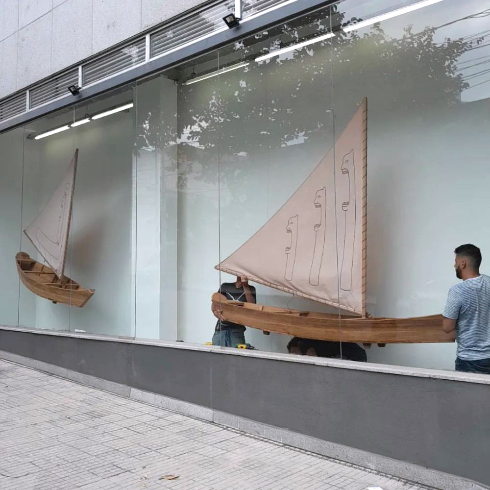 Vista externa da vitrine da galeria durante a montagem, com duas embarcações de madeira suspensas atrás do vidro. Técnicos ajustam a instalação no interior, enquanto a rua e árvores se refletem na fachada.