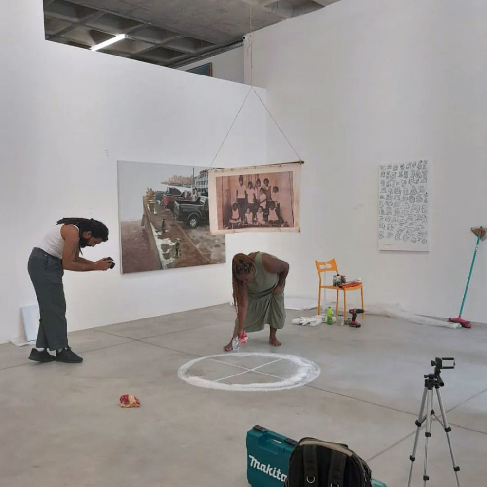Registro de montagem da exposição maa: artista desenha um círculo com pó branco no chão da galeria enquanto outra pessoa fotografa o processo. Ao fundo, obras já instaladas nas paredes e materiais de montagem espalhados pelo espaço.