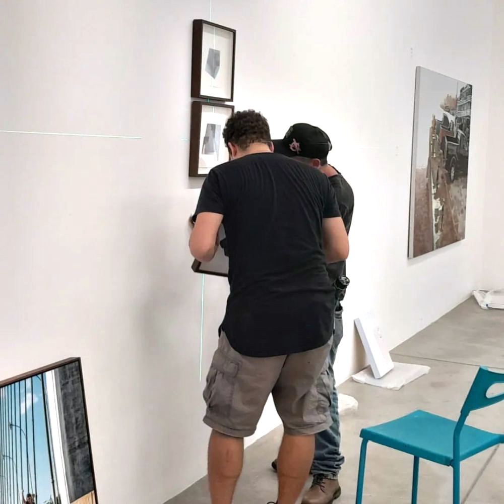 Dois montadores alinham quadros em uma parede branca utilizando nível a laser verde. Outras obras já estão instaladas ao redor, e ferramentas permanecem apoiadas no chão durante o processo de organização.