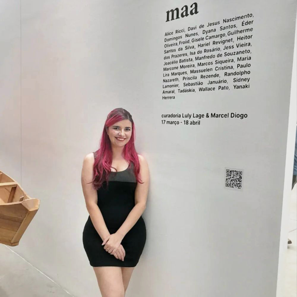 Retrato da curadora Luly Lage ao lado do texto de parede da exposição maa, com lista de artistas participantes e datas. Ela veste vestido preto e sorri para a câmera dentro do espaço expositivo.