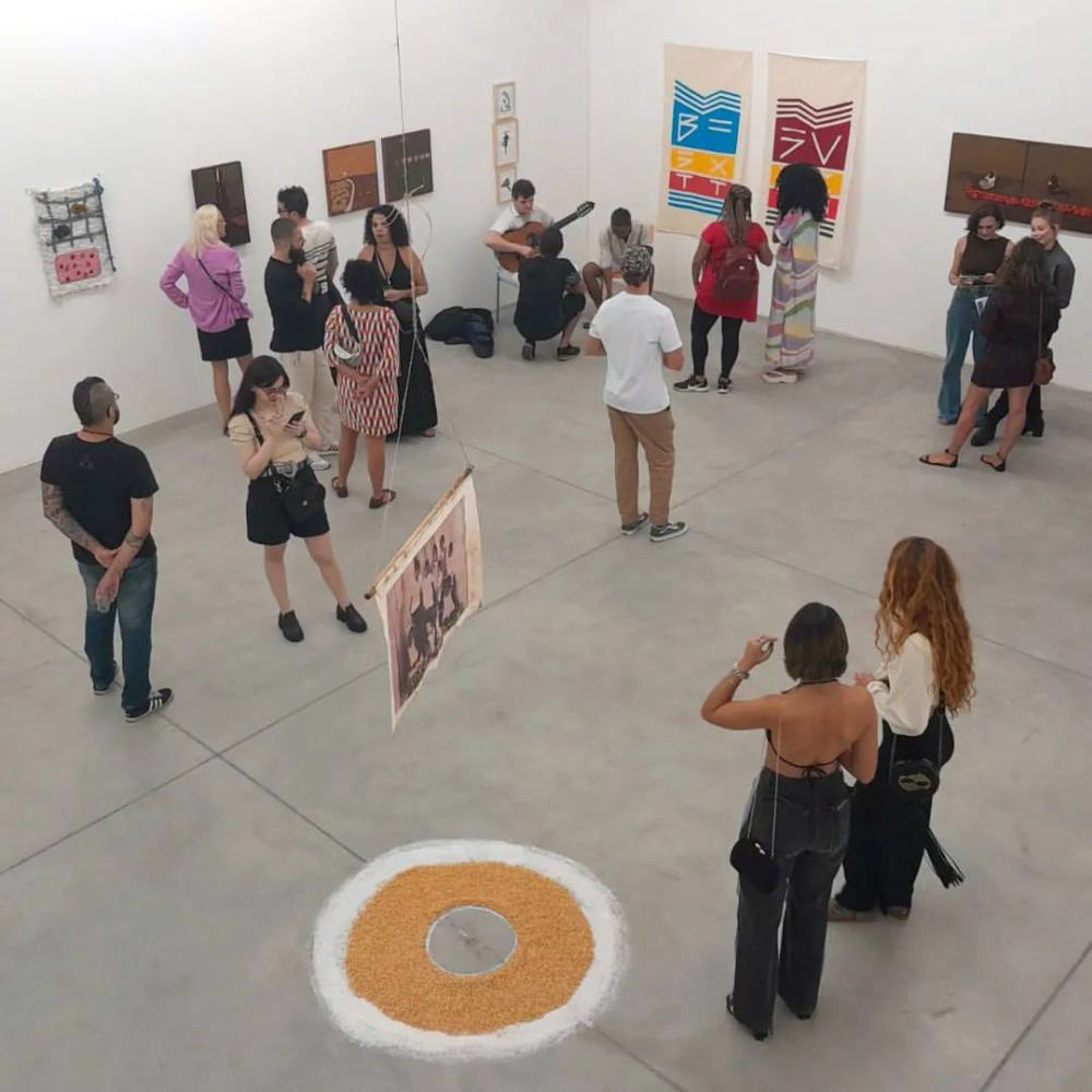 Vista aérea da abertura da exposição maa, com visitantes circulando pelo salão branco da galeria. No centro do chão, uma instalação circular em grãos claros delimita a obra, enquanto pessoas conversam e observam trabalhos nas paredes.