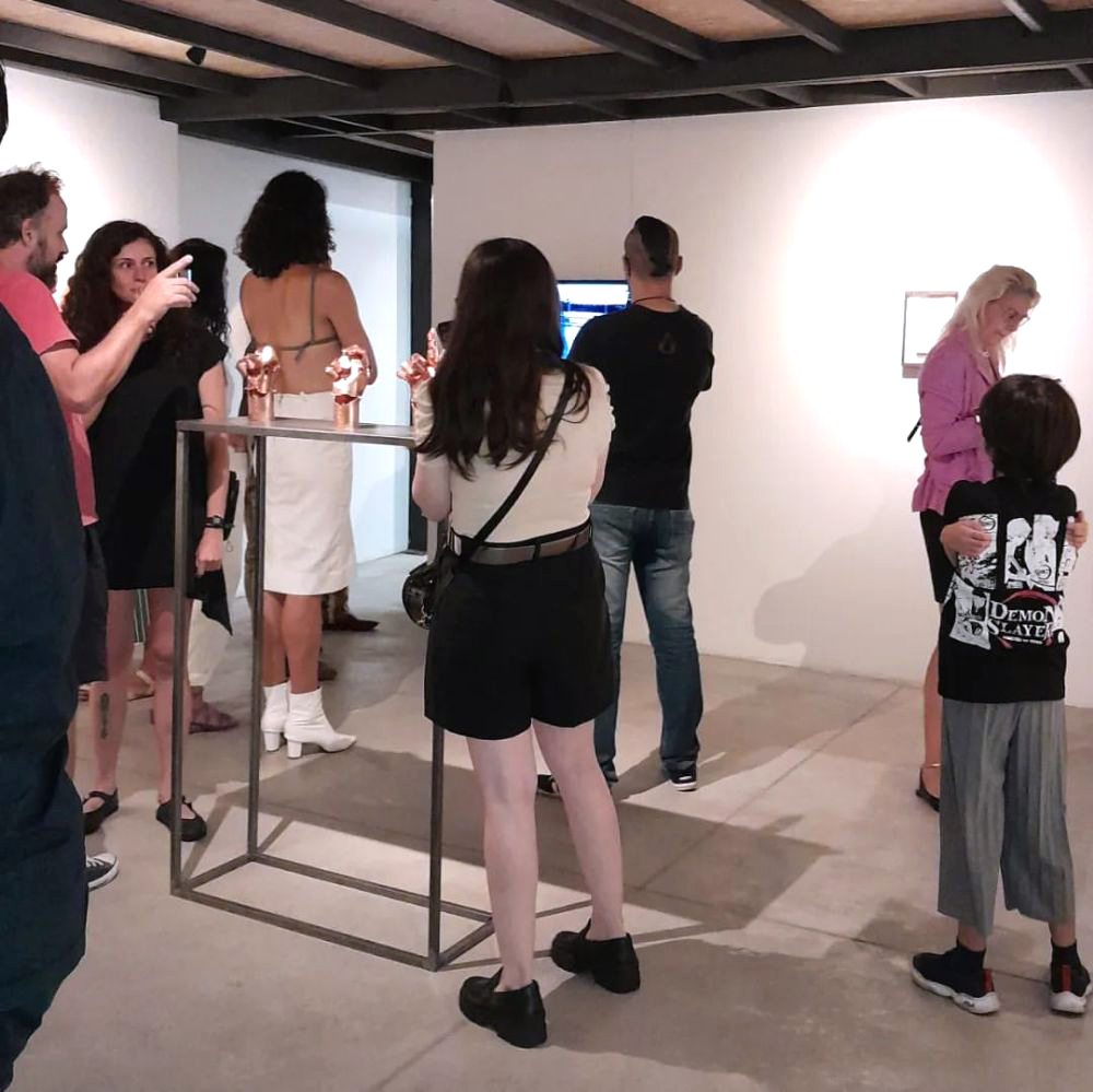 Público reunido em frente a obras na parede durante a abertura da exposição. Um grupo conversa próximo a um pedestal metálico com pequenos objetos expostos, enquanto outros visitantes exploram o espaço.