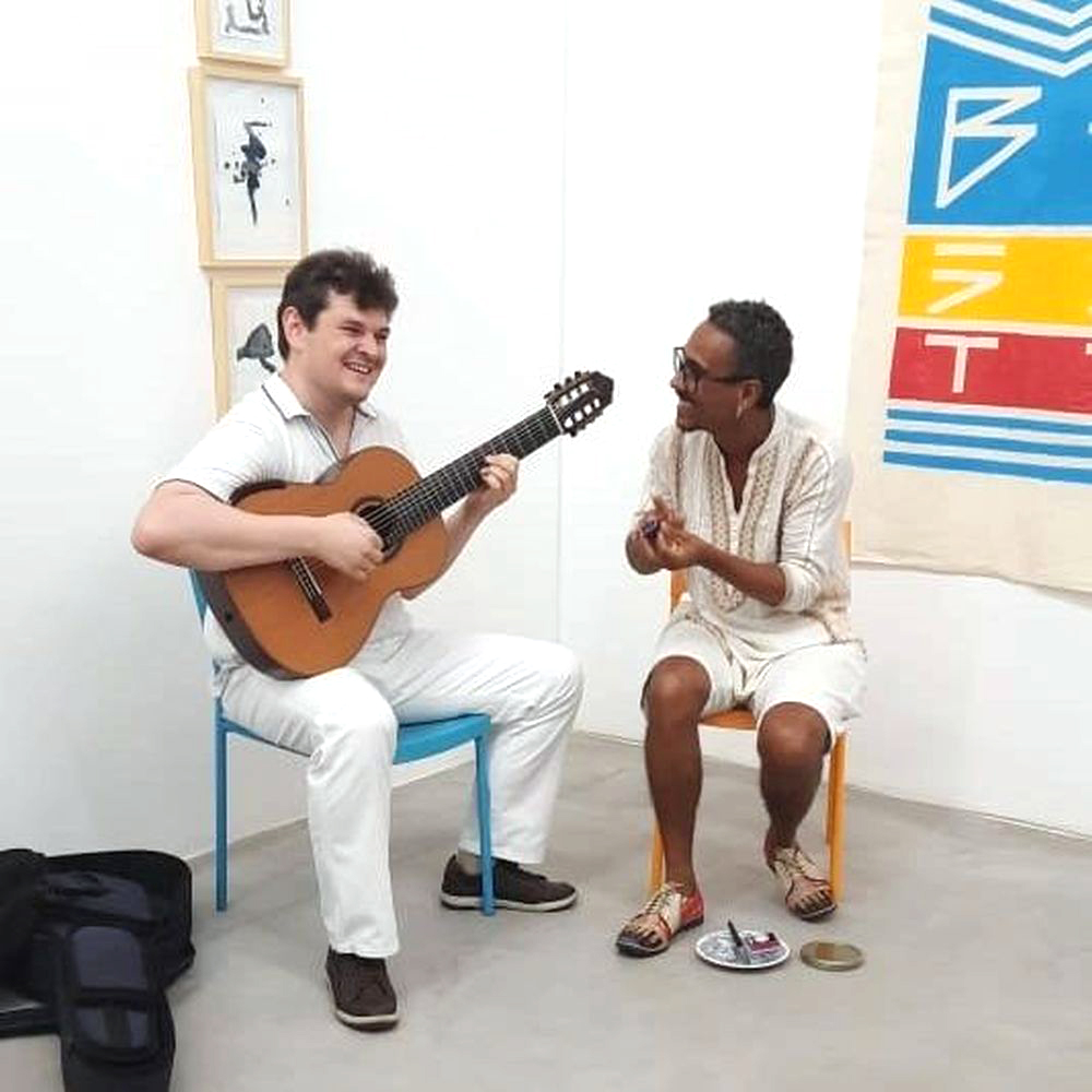 Dois músicos sentados em cadeiras coloridas realizam apresentação ao vivo dentro da galeria: um toca violão enquanto o outro canta e marca o ritmo com as mãos, criando atmosfera intimista durante o evento.