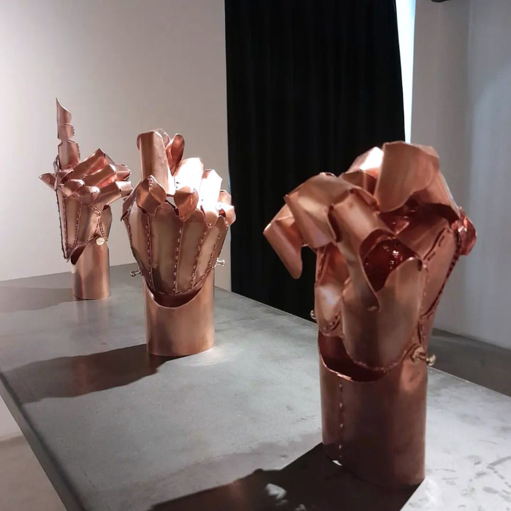Três esculturas metálicas em tom cobre dispostas sobre um grande pedestal cinza. As formas lembram mãos ou estruturas orgânicas abertas, com placas dobradas e rebites aparentes, iluminadas lateralmente contra fundo branco e cortina preta.