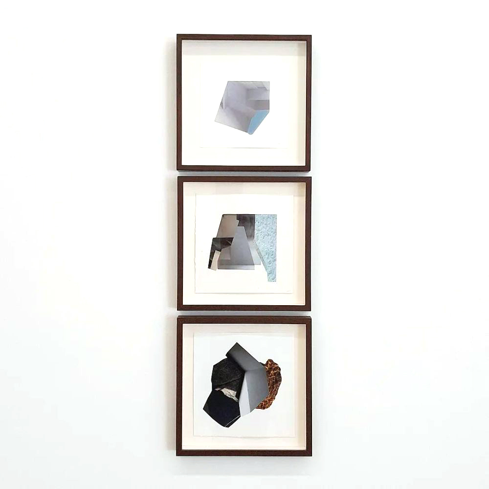 Trio de obras geométricas emolduradas, instaladas verticalmente na parede branca. Cada composição apresenta formas recortadas em tons de cinza, preto e azul, criando volumes abstratos e sobreposições sutis dentro de molduras de madeira escura.