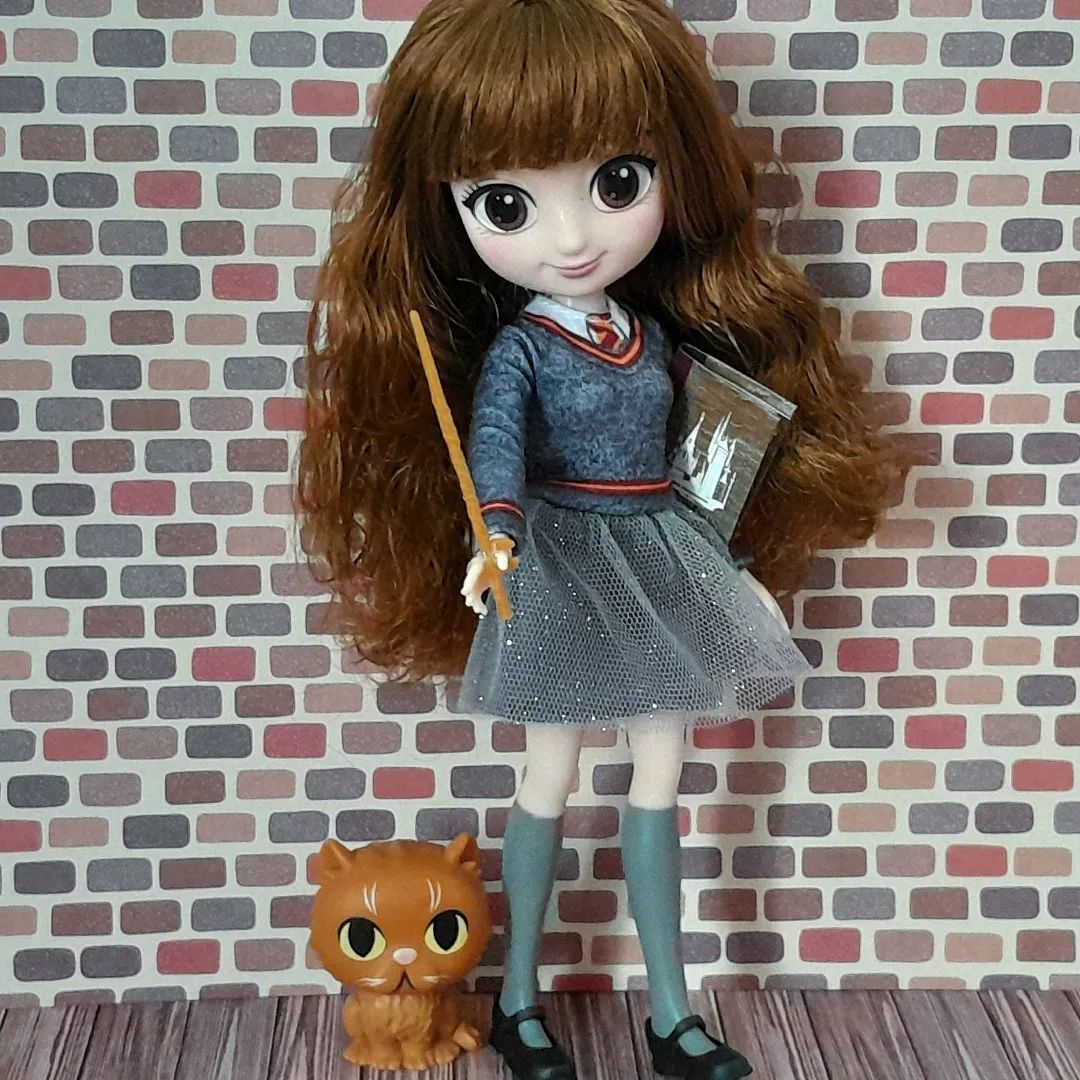 Boneca representando a personagem Hermione Granger, com cabelos castanhos e longos, pele clara, uniforme cinza da Escola de Hogwarts e uma varinha na mão