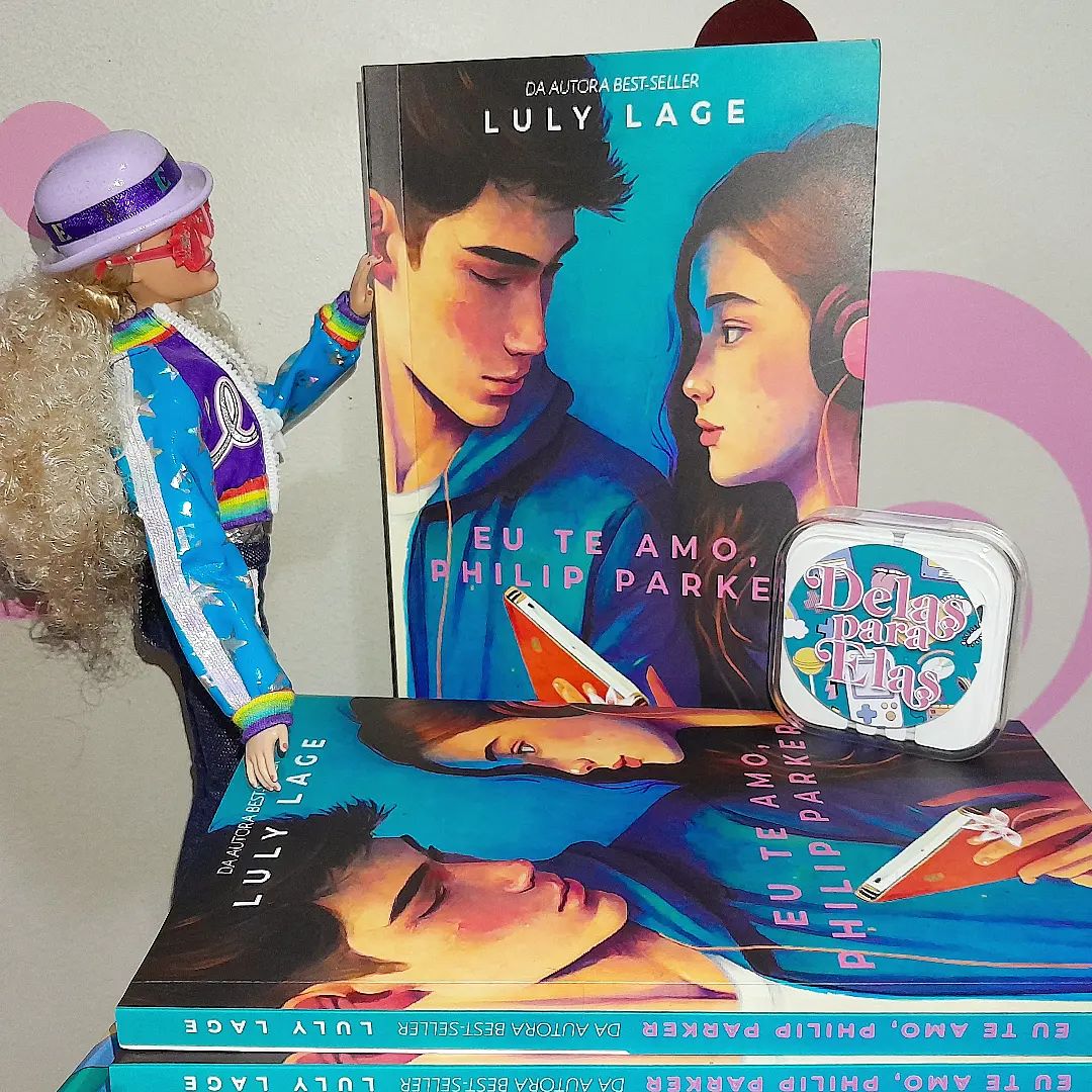 Cópias físicas do livro EU TE AMO, PHILIP PARKER, com capa turquesa e ilustração de um jovem casal olhando um para o outro, com o título abaixo. Ao lado dos livros há uma boneca Barbie temática do cantor Elton John, com cabelos loiros cacheados e óculos de sol cor de rosa, posando de lado como se olhasse para os livros.