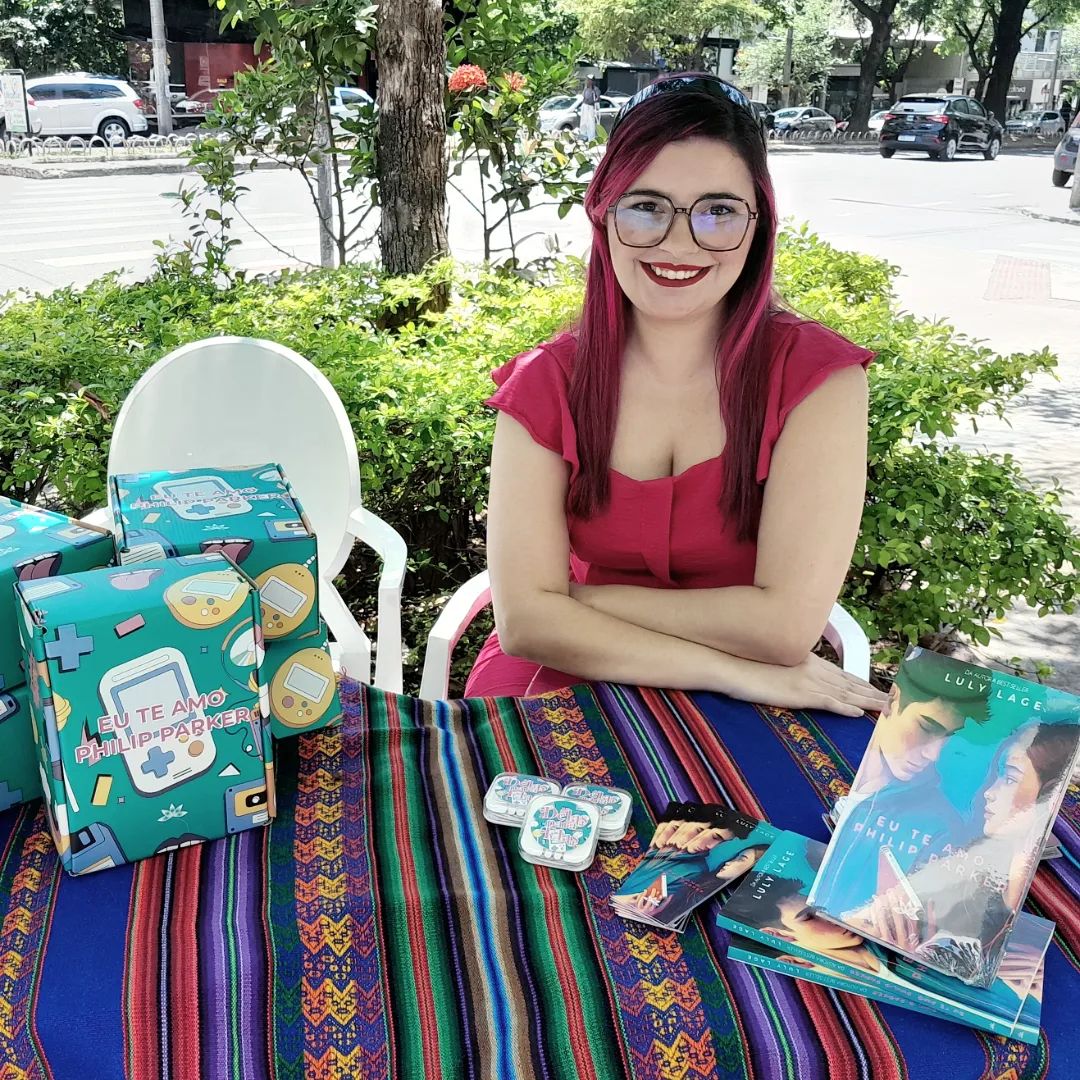 Foto de Luly Lage, com cabelos rosa e óculos de grau marrom escuro, vestindo um macacão rosa choque sentada atrás de uma mesa onde há edições do livro EU TE AMO, PHILIP PARKER expostas.