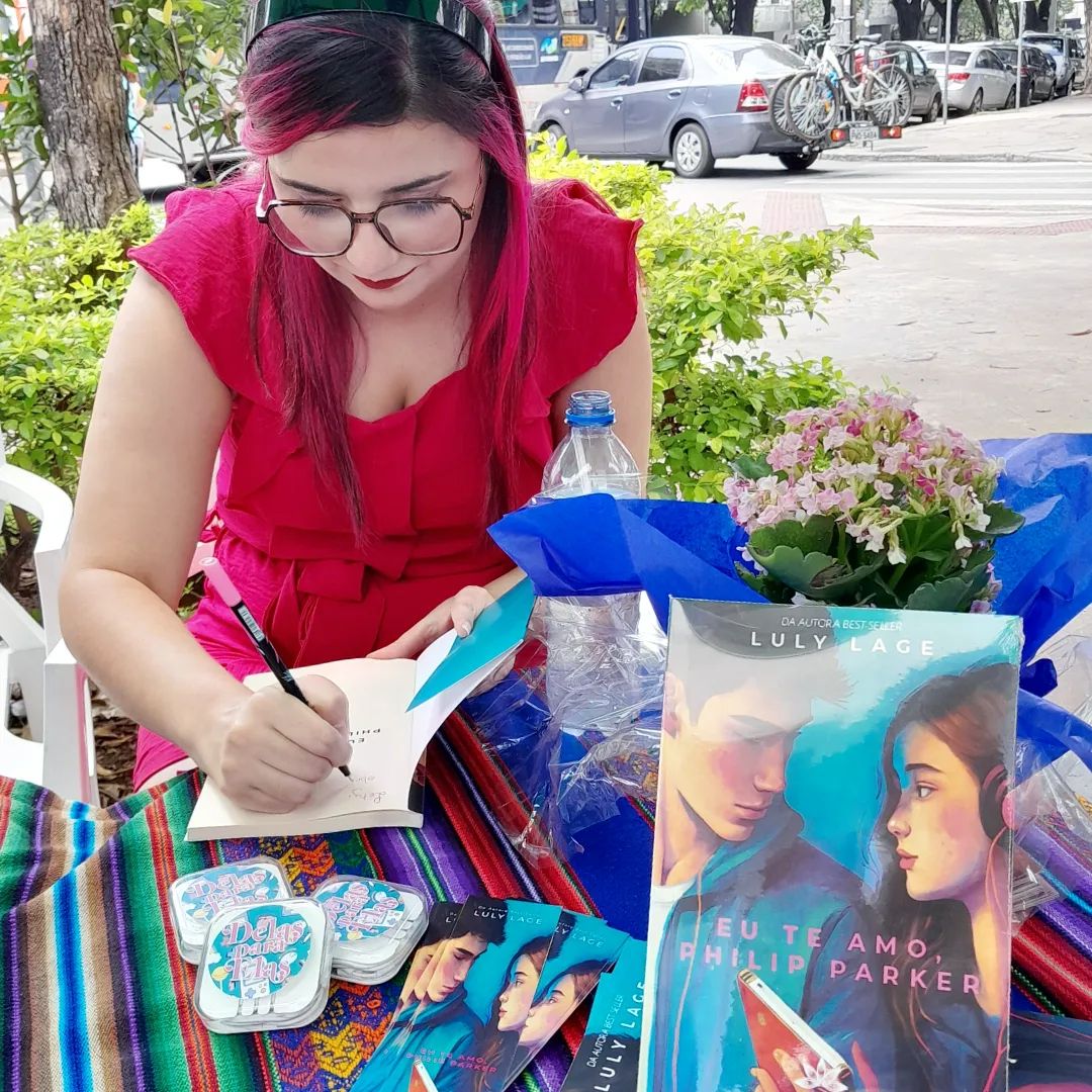 Foto de Luly Lage, com cabelos rosa e óculos de grau marrom escuro, vestindo um macacão rosa choque sentada atrás de uma mesa autografando edições do livro.