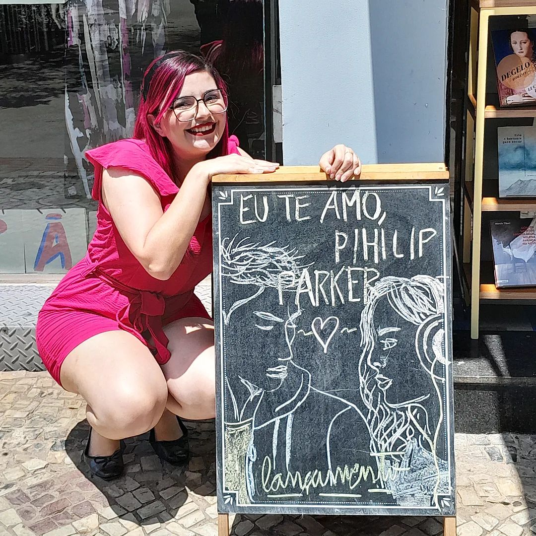Foto de Luly Lage, com cabelos rosa e óculos de grau marrom escuro, vestindo um macacão rosa choque agachada ao lado de um pequeno quadro negro onde há um desenho simulando a capa.