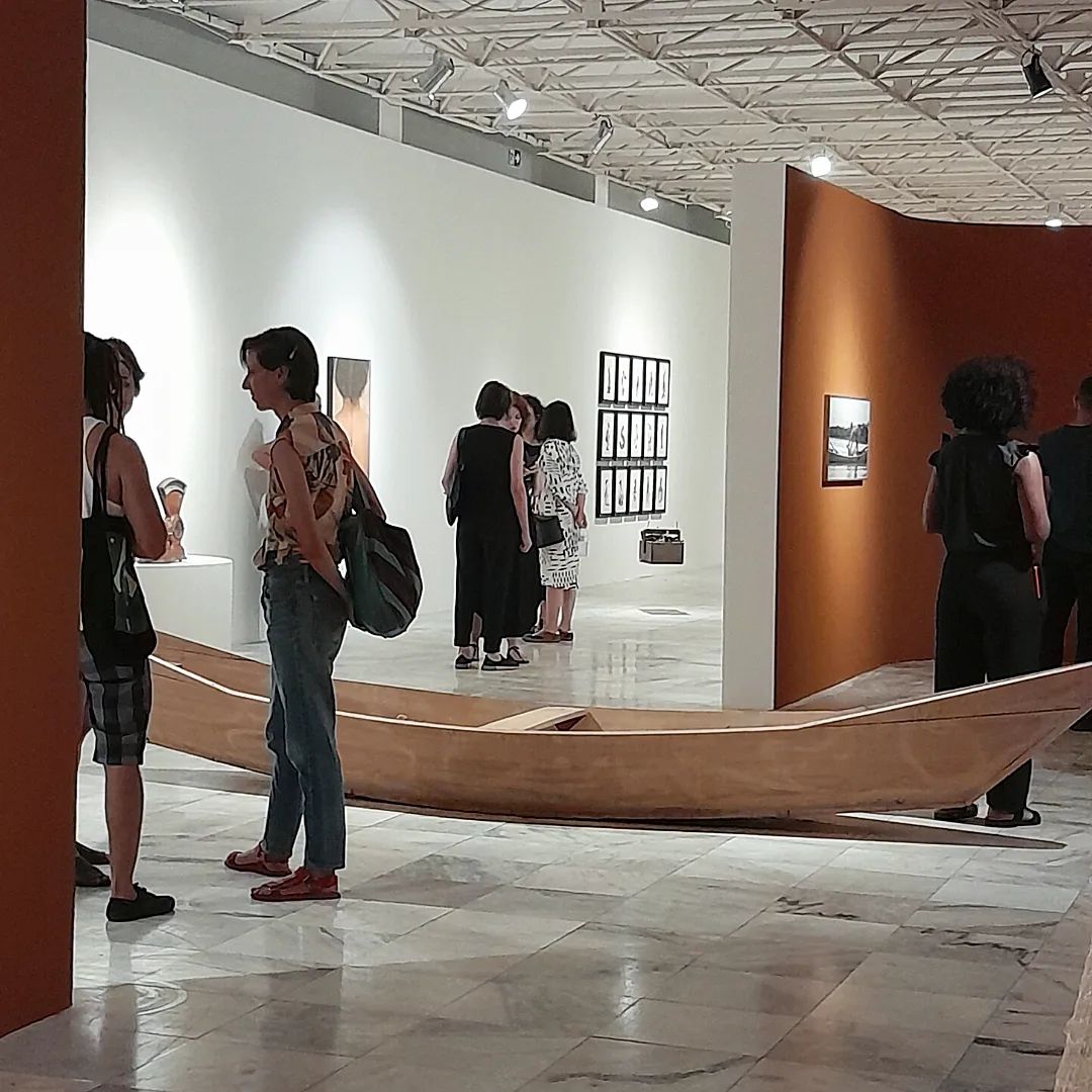 Vista da exposição madeira doce, água áspera, de davi de jesus do nascimento, com destaque para embarcação sem vela forrada de tamarindos. Ao redor, visitantes apreciam a área expositiva. Na lateral direita, existem fotografias em parede alaranjada e à esquerda, ao fundo, aquarelas em parede branca.
