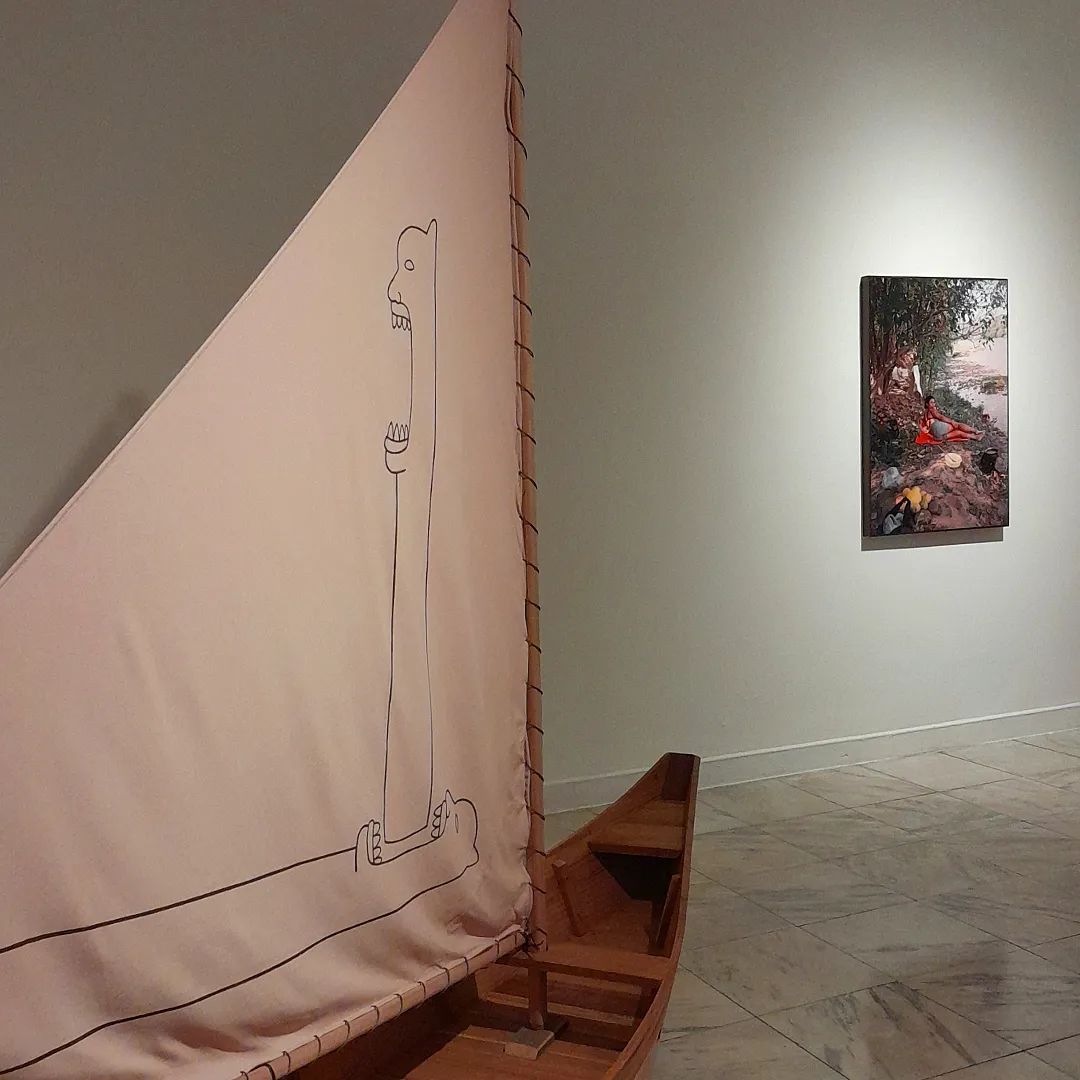 Embarcação de madeira com vela de tecido onde há um desenho de carranca. Ao fundo, uma fotografia de família do artista ampliada.
