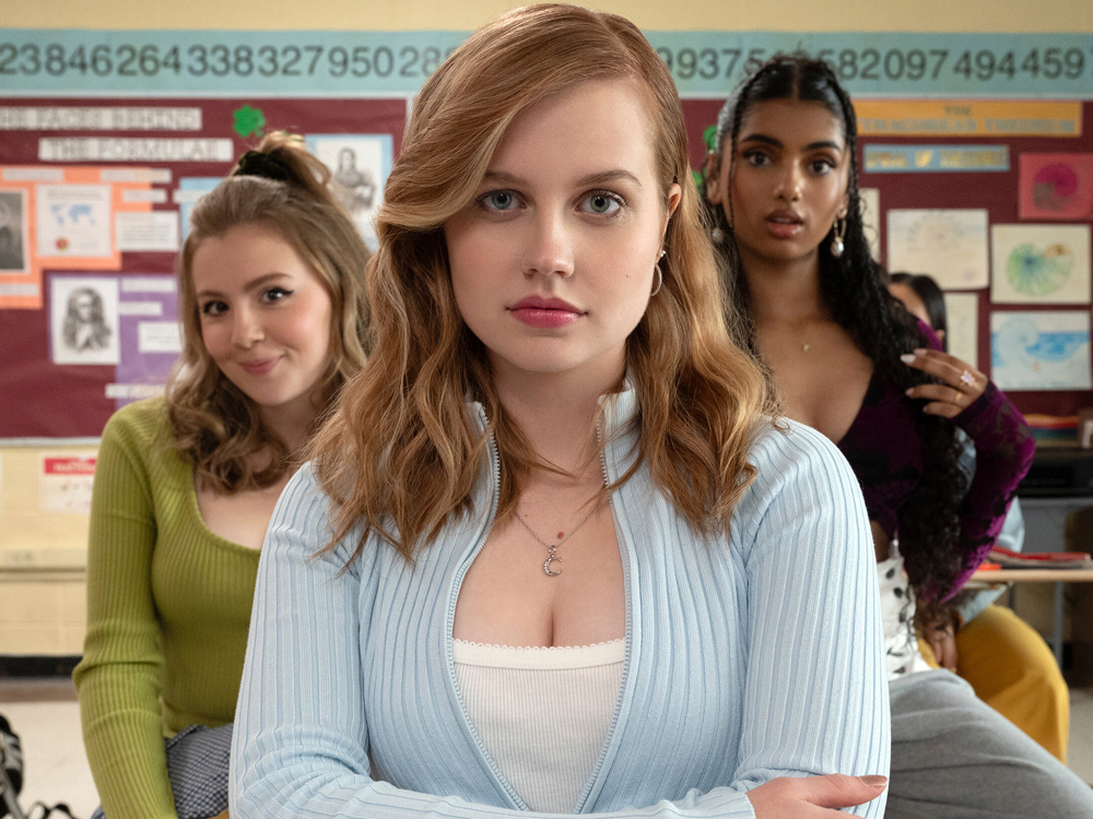 A protagonista de Meninas Malvadas, Cady, aparece em primeiro plano, de braços cruzados e expressão série. Atrás dela estão as patricinhas Cretchen e Karen, indicando que é a líder do trio.