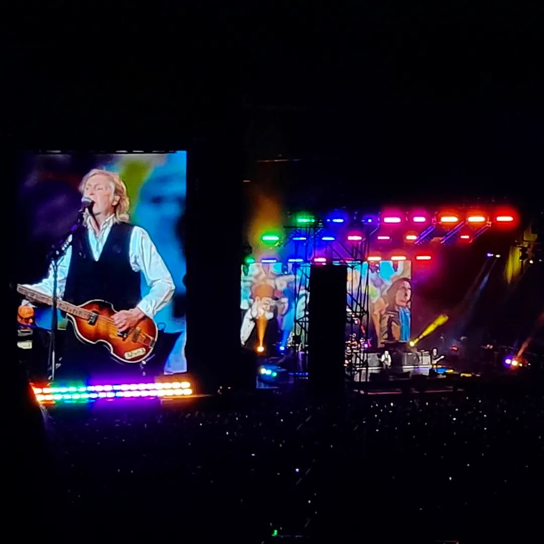 Momento do show em que o palco está iluminado com cores do arco-íris e Paul McCartney, além de cantar, toca baixo. Apenas um dos telões é visível na foto.