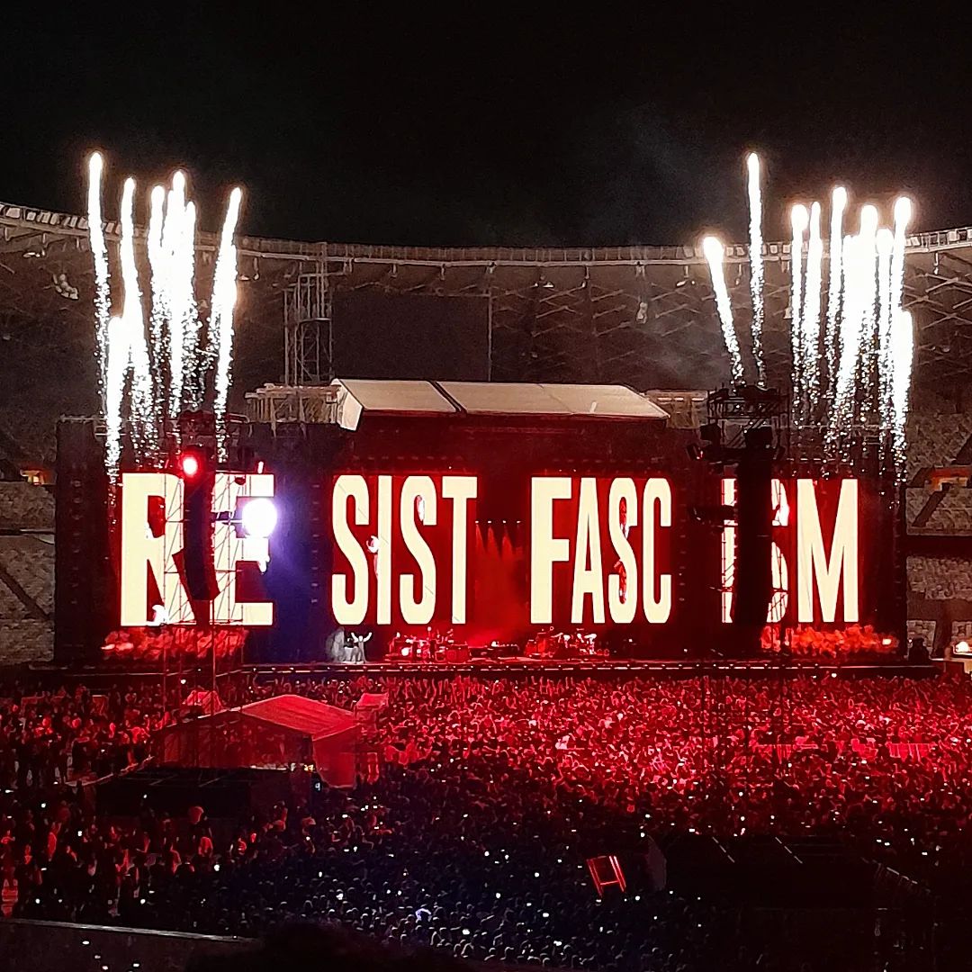 Palco do show visto da plateia, onde Roger Waters encontra-se ao centro, quanse imperceptível por causa da distância, e o telão atrás do mesmo está vermelho com os dizeres RESIST FASCISM.
