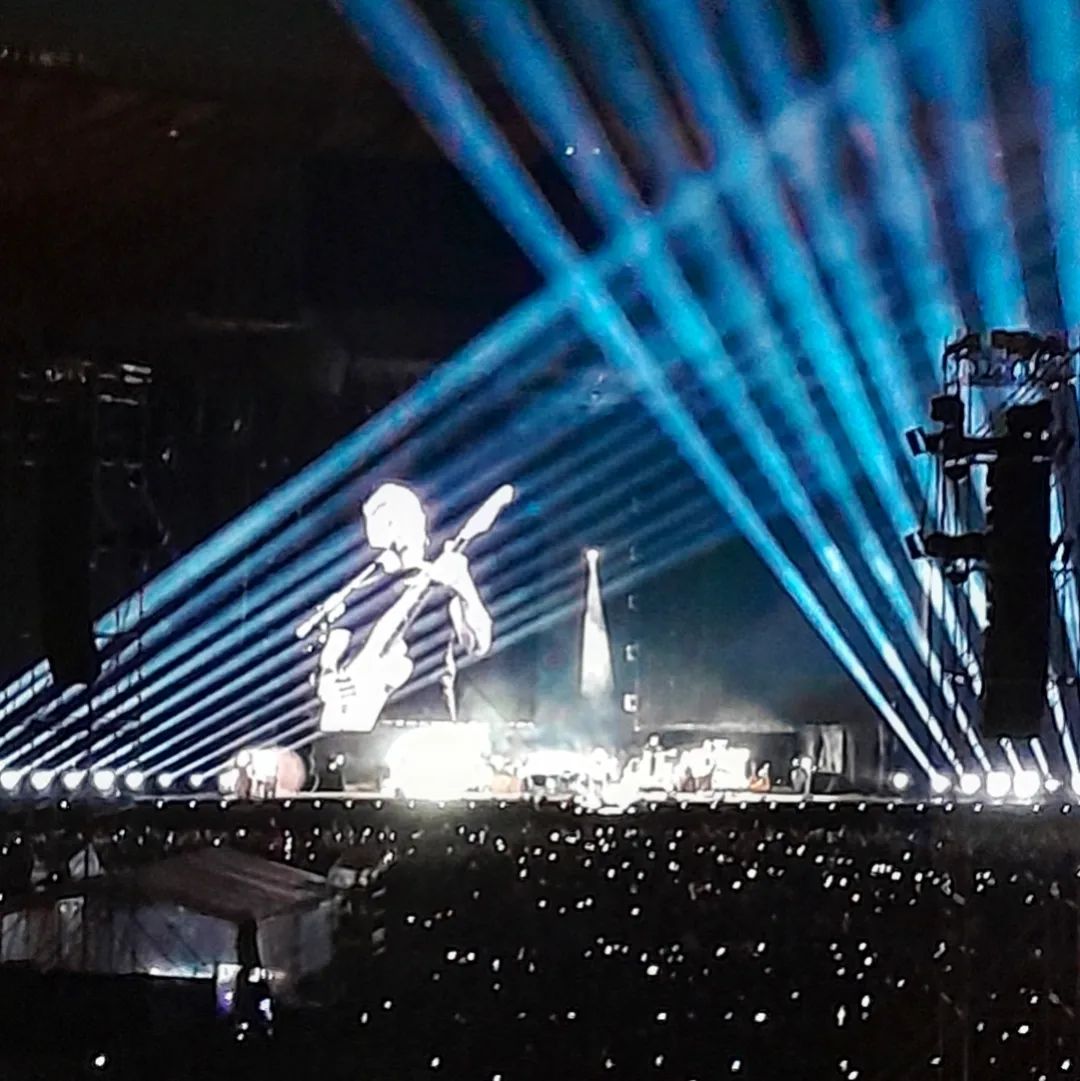 Foto que mostro Roger Waters no telão, com muitas luzes claras ao seu redor, tocando uma guitarra.