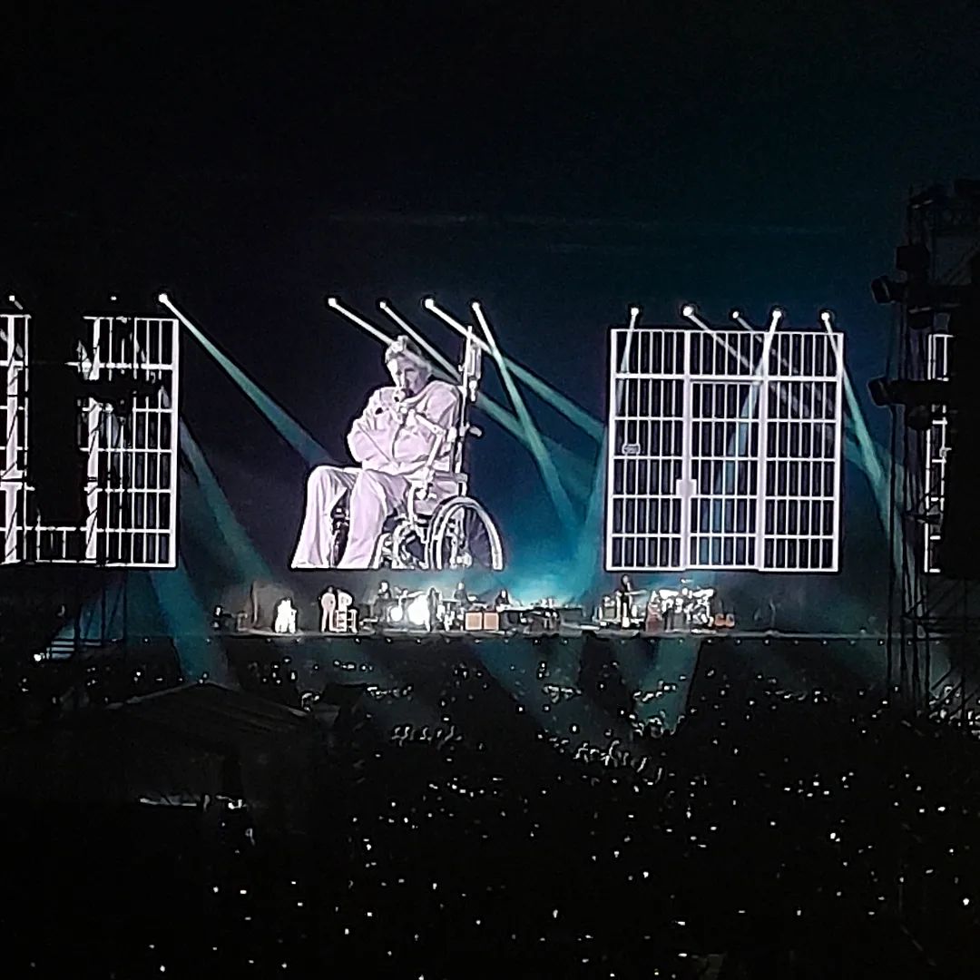 Foto do show em que Roger Waters aparece no telão, sendo visível em frente à sua imagem. Nesse momento, ele canta In The Flesh sentado em uma cadeira de rodas, amarrado em uma camisa de força.