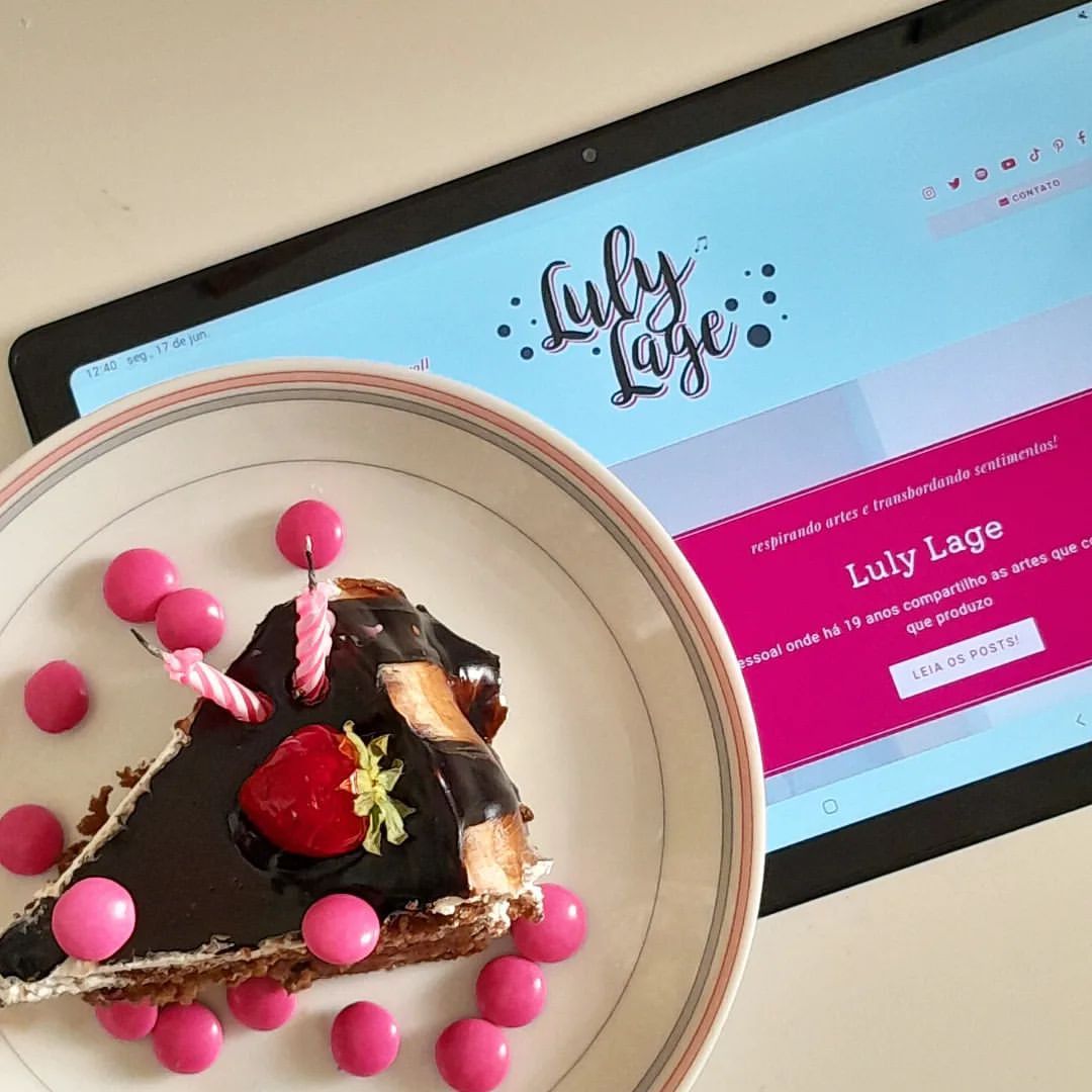 Foto vista de cima onde a página inicial do blog está aberta em um tablet e, sobre ele, há um bolo de chocolate com morango, confeito cor de rosa e duas velas apagadas sobre ele