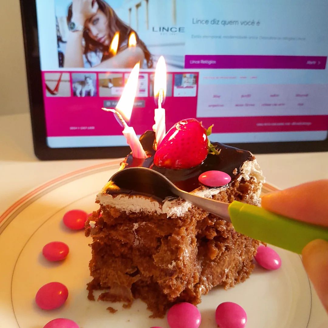 A tela do tablet agora mostra o rodapé da página de posts do blog,  as velas sobre o bolo estão acesas e há uma colher partindo o mesmo, que já teve um pedaço comido.