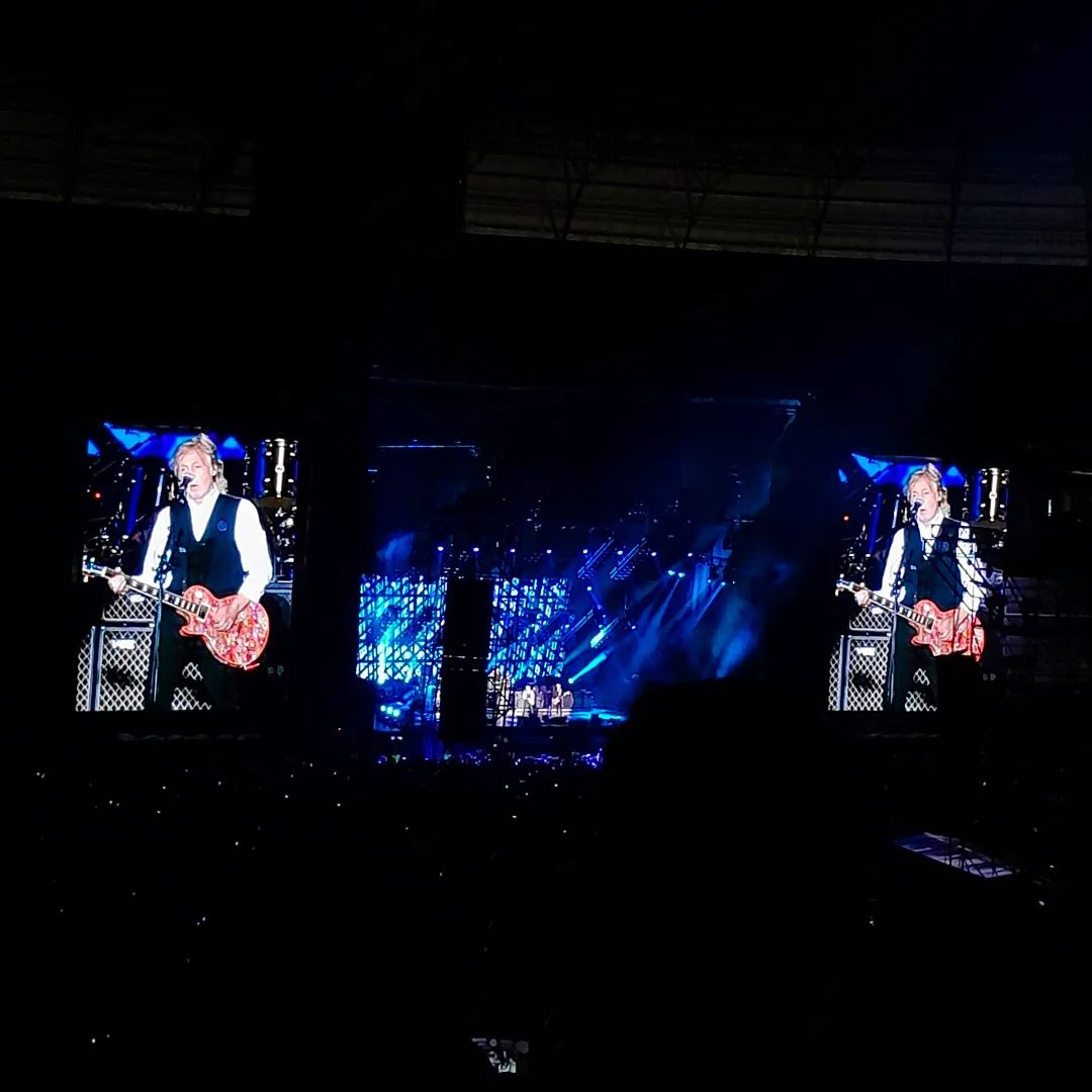 Imagem no mesmo ângulo da primeira, com iluminação do palco azulada. Nela, Paul toca um violão.