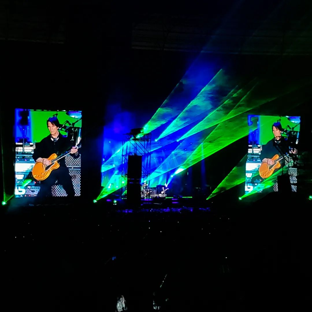 Vis]ao geral do palco, com iluminação azul e verde e um outro membro da banda tocando uma guitarra em destaque no telão.
