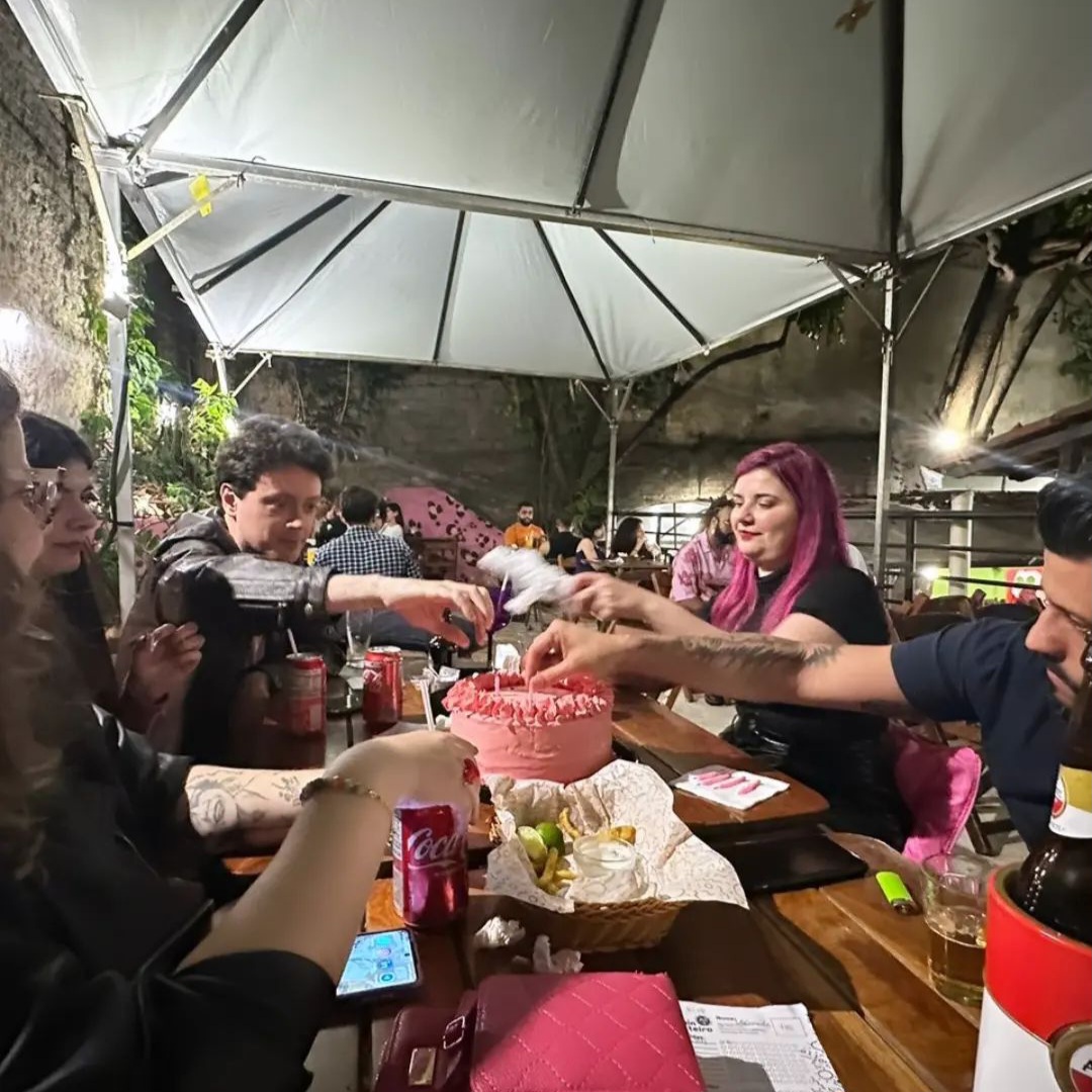 Foto da mesa de bar onde há um bolo cor-de-rosa no centro e as pessoas ao redor, dois homens e três mulheres, tiram velas da mesma cor de cima dele