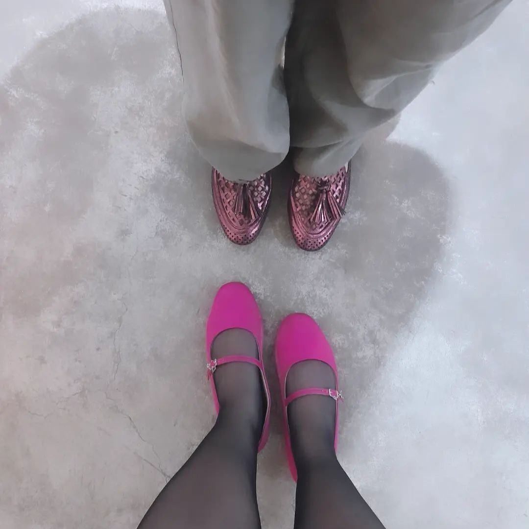 Dois pares de pernas femininas e seus pés, a primeira usando uma calça cinza e sapato rosa metálico, a outra usando meia calça preta e sapatilha rosa-choque