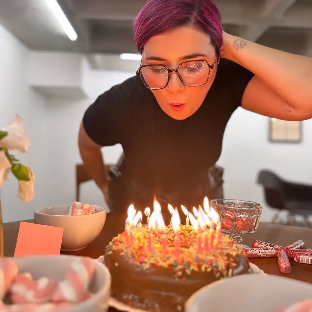 Foto onde estou curvada sobre outra mesa, dentro de um escritório, soprando velas de um bolo