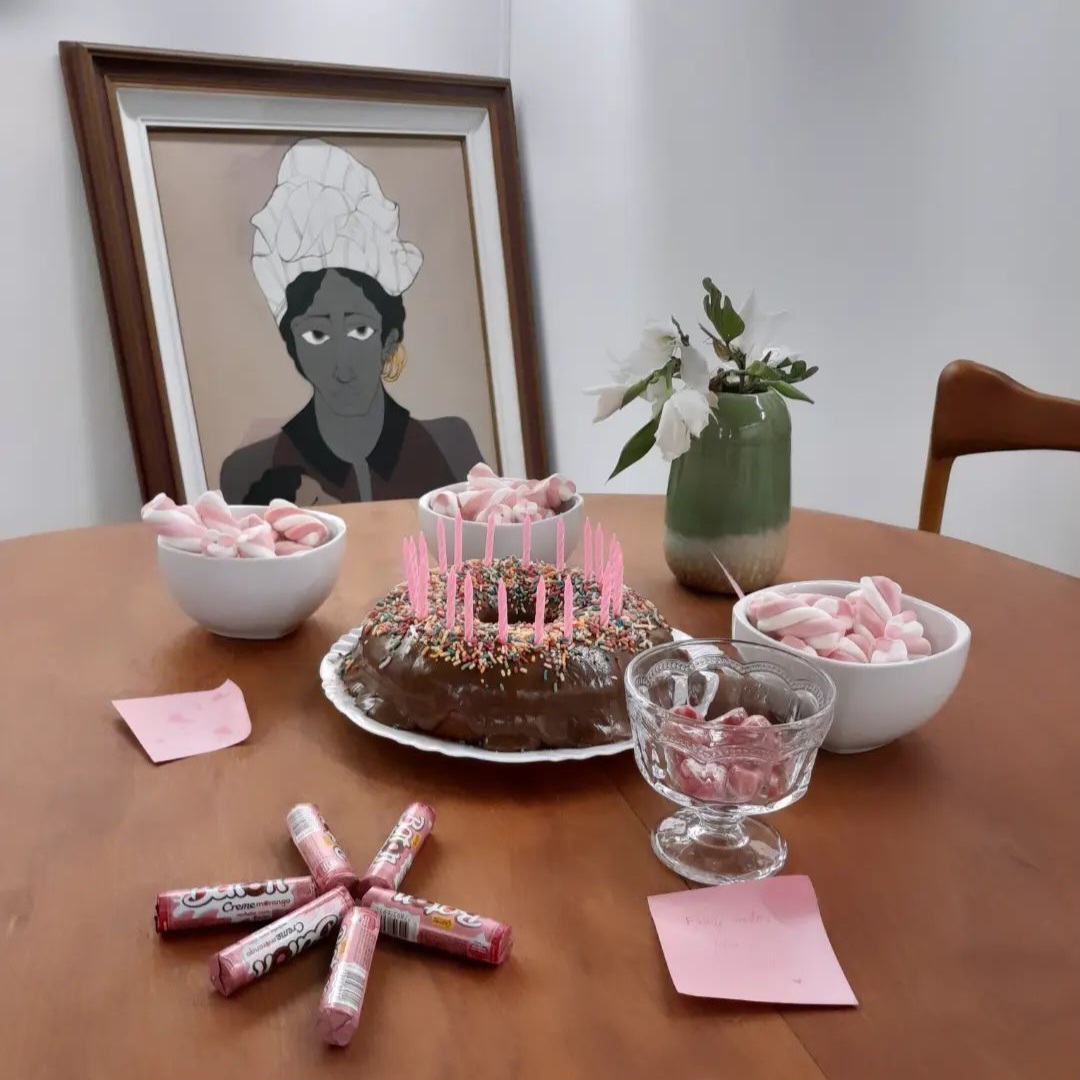 Foto da mesa da foto anterior, onde o bolo de chocolate está no centro com as velas ainda apagadas e outros doces cor de rosa, como balas, marshmallows e chocolates, estão ao seu redor